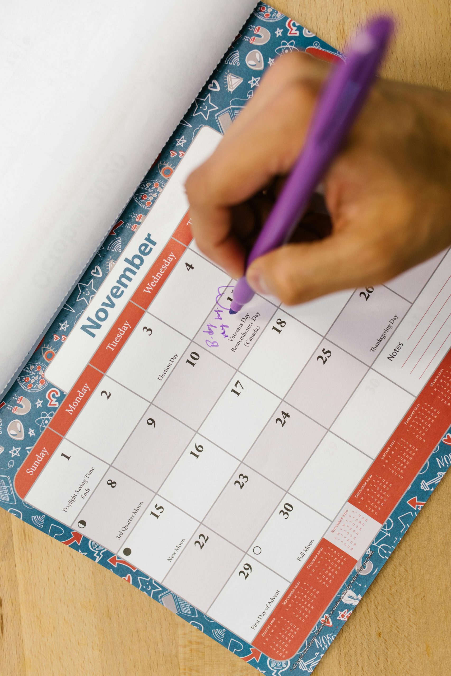 A person writes a reminder on a colorful November calendar using a purple pen.